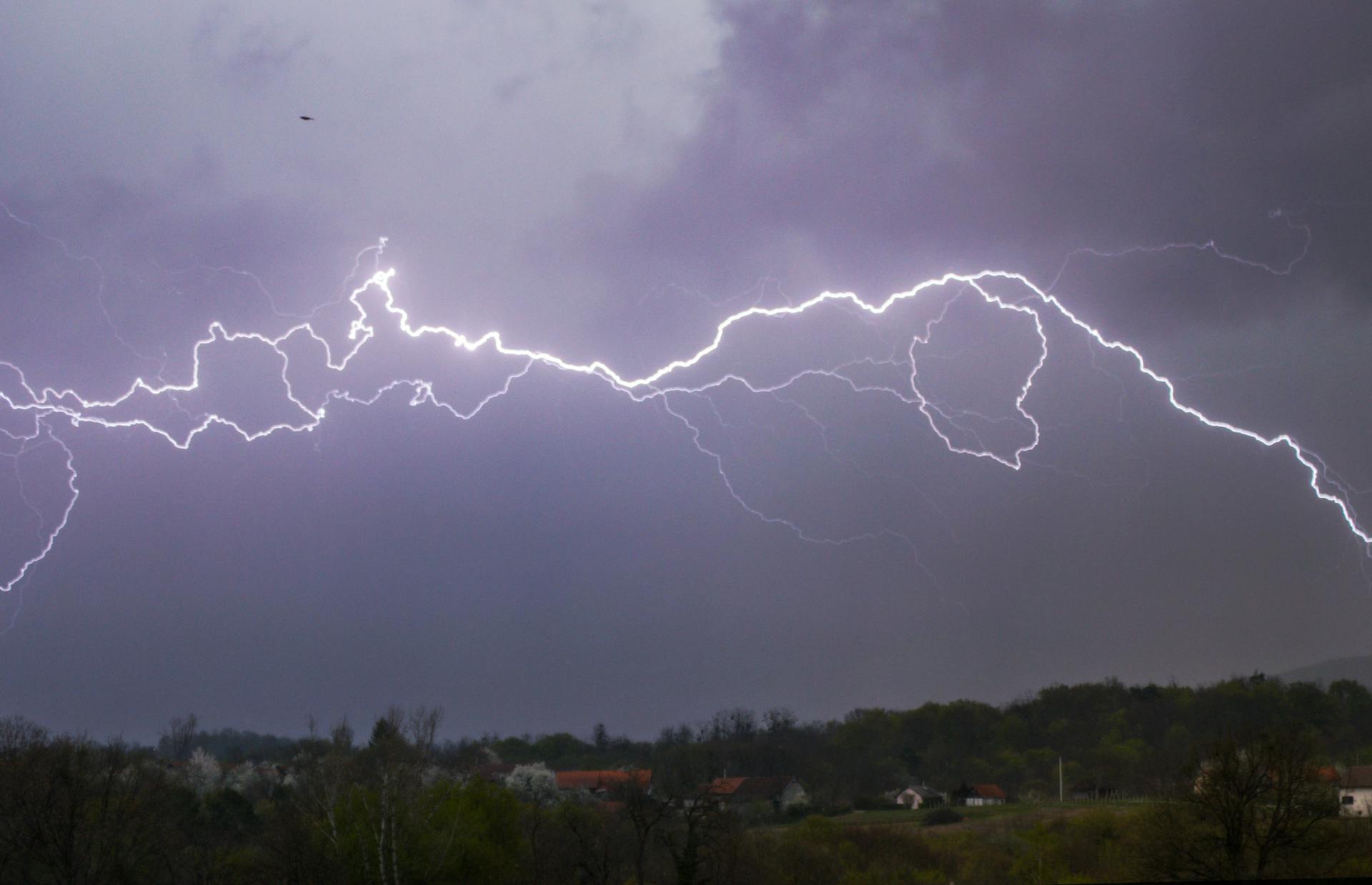 Hail and wind ratings for asphalt shingles illustrated by a bright lightning strike across dark storm clouds over a rural neighborhood.